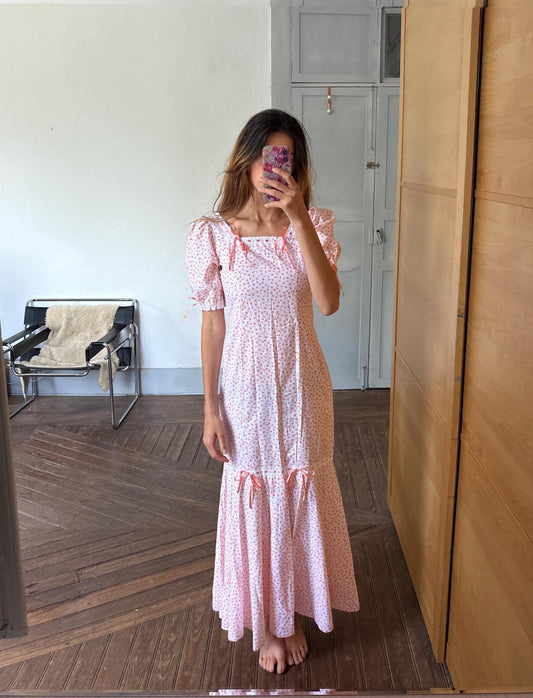 May include: A light pink dress with a floral pattern and a square neckline. The dress has short, puffed sleeves and is adorned with small pink bows. The skirt is floor-length. The dress is made of a light fabric.