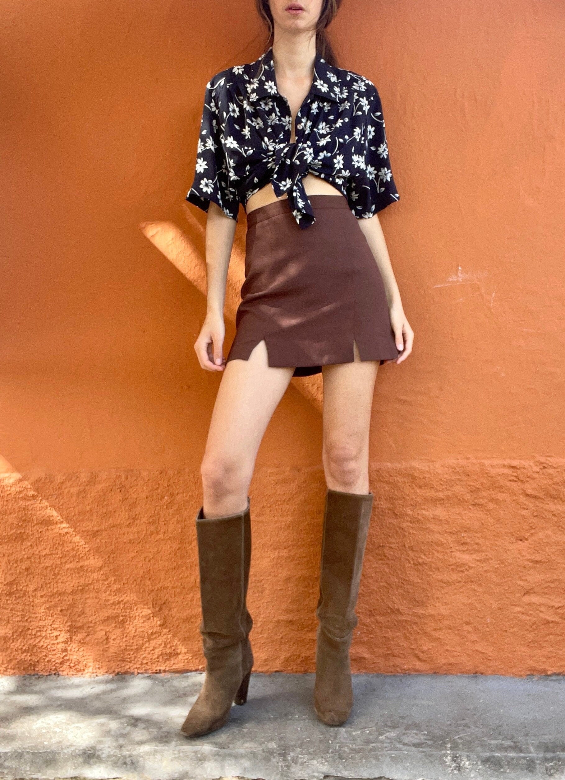 May include: A woman's outfit comprising a black floral print top, a brown mini skirt with a front slit, and brown suede knee-high boots. The outfit is set against an orange wall.
