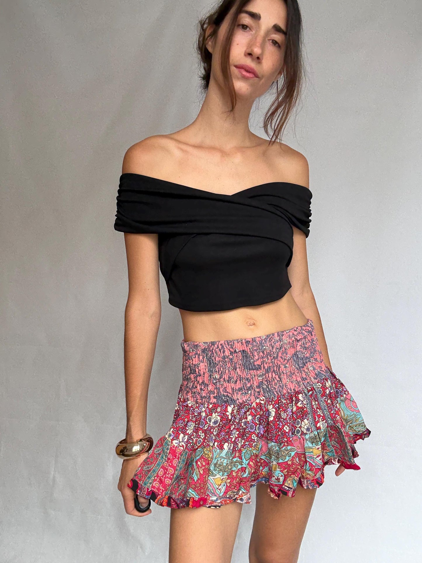 May include: A black, off-the-shoulder top and a patterned mini skirt. The skirt is a mix of pink, red, and teal floral and paisley designs. A gold bracelet adds a touch of elegance to the outfit. The skirt is approximately 30 cm in length.