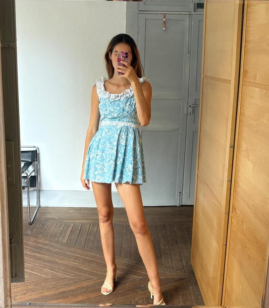 May include: A light blue floral print dress with white lace trim and ruffled edges. The dress has a sleeveless design and a short skirt. The person is wearing beige heels. The dress is reflected in a mirror.